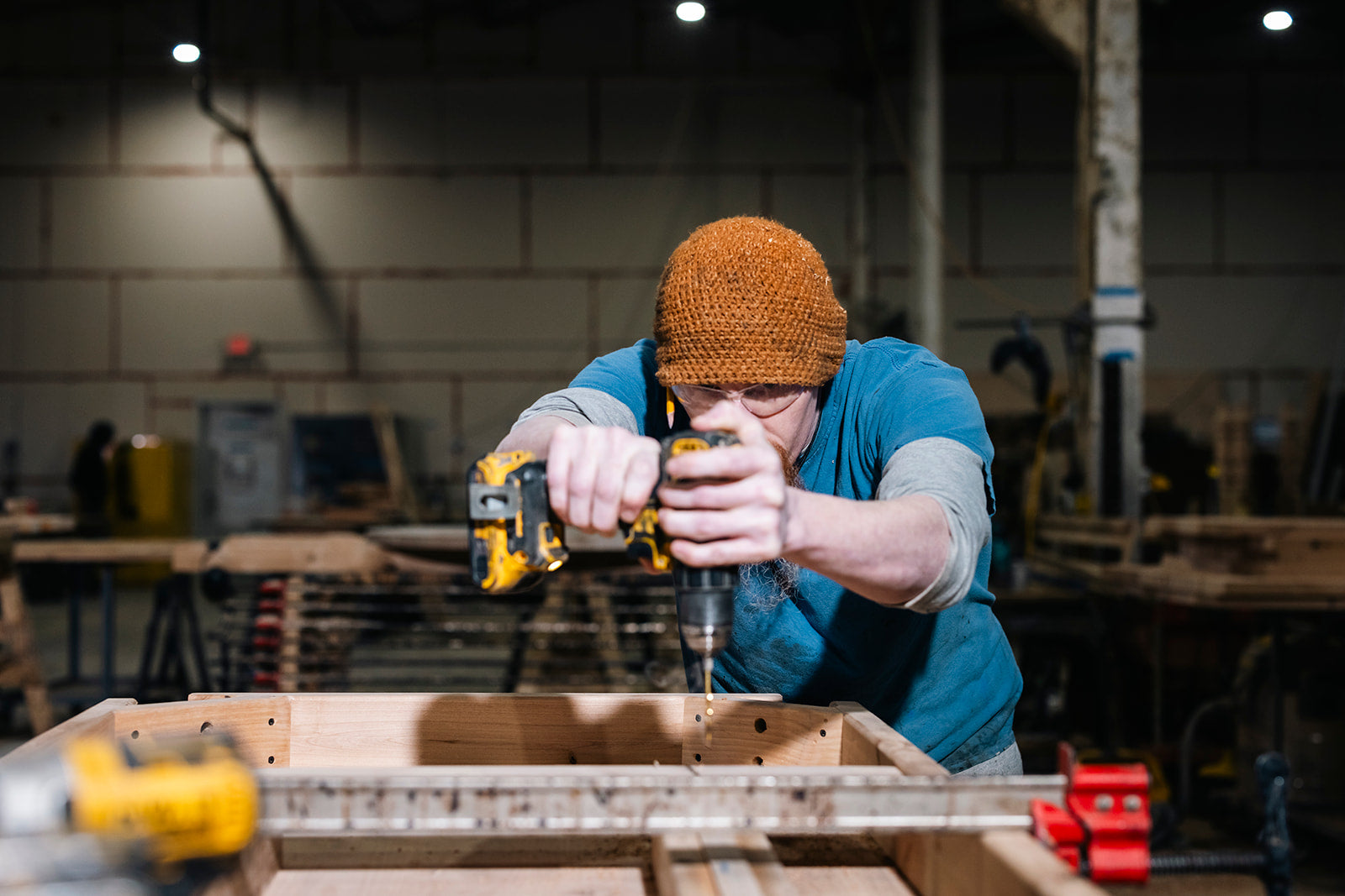 A person wearing a knitted hat and blue shirt uses a cordless drill on a wooden structure in a workshop. The background shows industrial tools and materials, with overhead lighting illuminating the workspace.