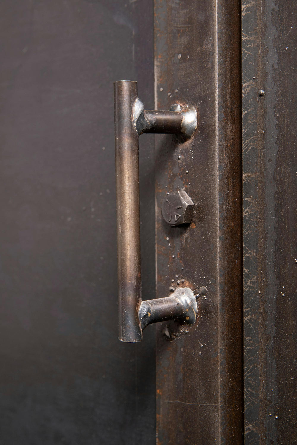Close-up of an industrial metal door handle attached to a dark, rusted surface. The handle is simple and cylindrical, with visible weld marks. A large bolt and a partially visible hexagonal fastener are nearby.