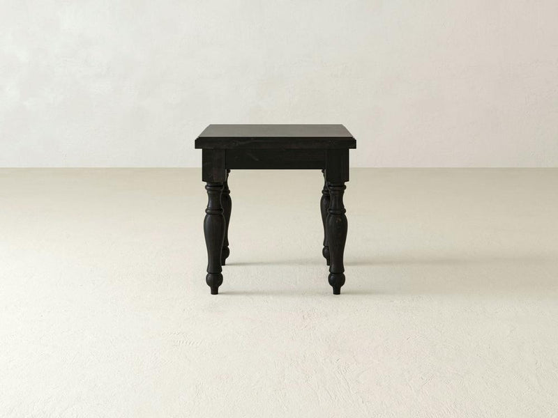 The Abigail End Table - Charred Ember by James+James, featuring ornate, turned legs in a black finish, stands on a plain, light-colored floor against a white wall. The table boasts a classic, vintage design with a smooth, rectangular surface.