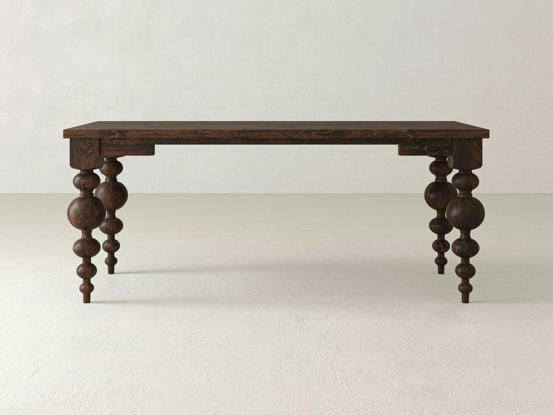 A dark wooden Olivia Desk from James+James, crafted from Knotty Alder, stands on a light, neutral-colored floor. The rectangular top is supported by four intricately carved Olivia Turned Legs, featuring a mix of spherical and cylindrical shapes stacked along their length.