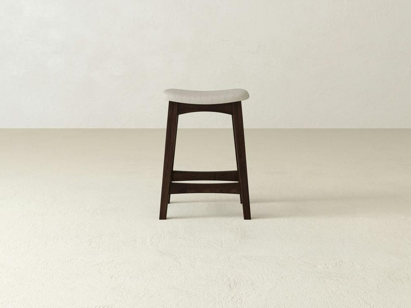 The James & James Weston Upholstered Stool, made from sturdy maple hardwood, features a minimalist design with clean lines and a dark finish. It has a light gray upholstered seat and is centered against a plain, light-colored wall and floor.