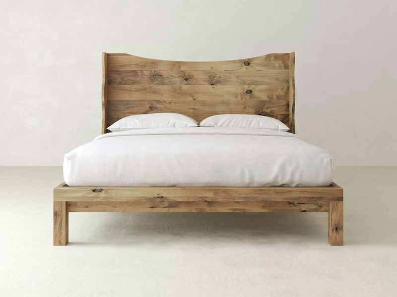 The James+James Harlow Bed exudes natural beauty with its solid wooden frame and headboard in a natural wood finish. This elegant wingback design, adorned with white bedding and two pillows, sits against a light-colored wall in a minimalist room, highlighting the simplicity and quality materials.