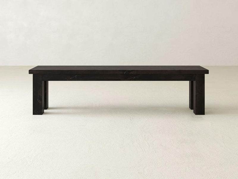 The James+James Farmhouse Bench in Charred Ember on Alder features clean lines and a dark wood finish, positioned on a light floor against an off-white wall.