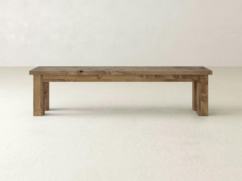 The James+James Farmhouse Bench in Harvest Wheat on Alder features straight legs and a rectangular seat, standing on a light, textured floor against a plain off-white wall.
