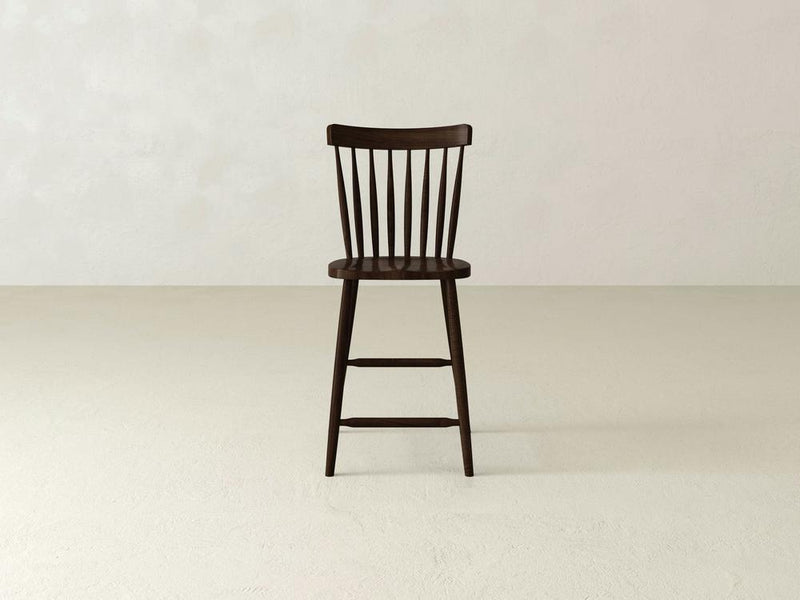 A Rustic Windsor Stool by James & James, made of durable maple hardwood, boasts a dark wooden spindle backrest. It stands on a light-colored floor in an empty room with textured white walls.