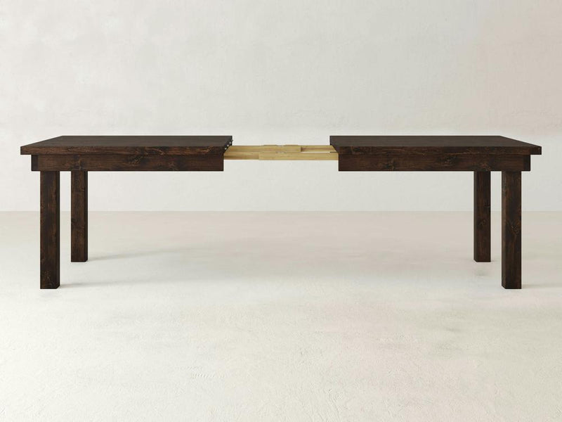 The Farmhouse Expandable Dining Table - Tobacco by James+James is shown in an expanded state, revealing its extension mechanism in the middle. The table features four sturdy legs and a simple, minimalist design. The plain, light-colored background enhances the dark wood's details and highlights the table's features.