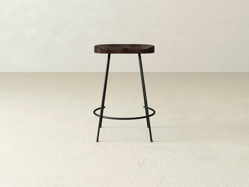 The Bryer Stool by James & James offers rustic charm with a minimalist wooden design, featuring a dark round seat and thin black metal legs. Its circular footrest adds an industrial touch, especially on light floors against off-white walls.