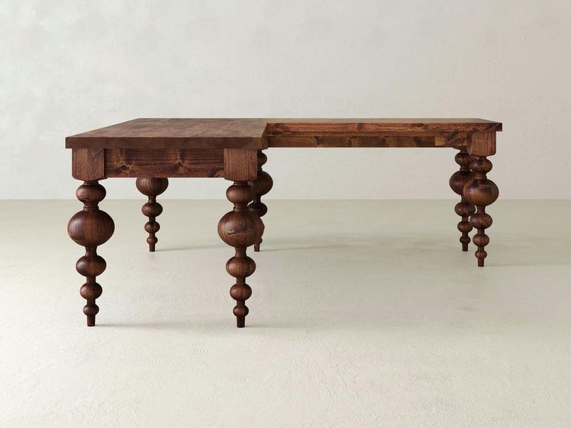 The Olivia Corner Desk by James+James is a wooden table with a distinctive design, featuring four intricately carved legs in a bulbous pattern. The handcrafted hardwood top has a smooth finish, and the right half is at a slightly lower height than the left, giving it an asymmetrical appearance. It makes for an ideal home office centerpiece.