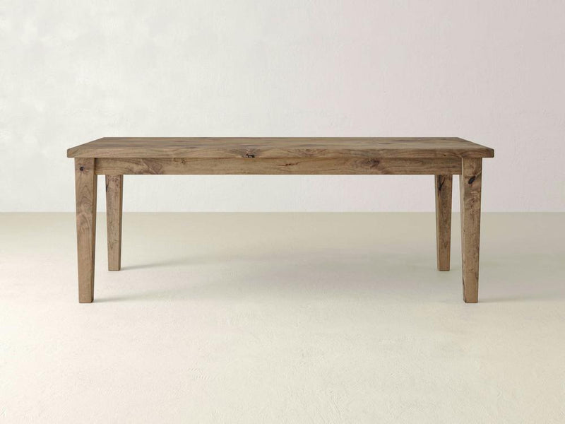 An Ava Parsons Dining Table - Harvest Wheat from James+James stands on a light-colored floor against a pale wall. The simple, rectangular wooden dining table features straight legs and a smooth top, showcasing the natural grain and texture of the wood with its natural finish.
