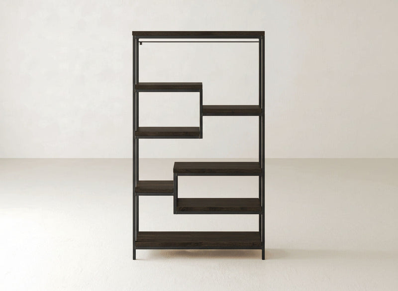 The James+James Arlo Shelf is a modern bookshelf with a geometric, zigzag design. It features multiple solid hardwood shelves framed in industrial steel, creating open spaces of varying heights and widths that are perfect for books or decor. The background consists of plain, light-colored walls and floors.