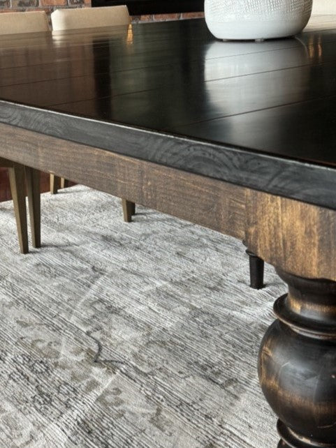 A wooden dining table with a dark polished surface and thick, rounded legs stands on a textured gray and white rug. A white vase is partially visible on the tables surface. Chairs with wooden legs are in the background.