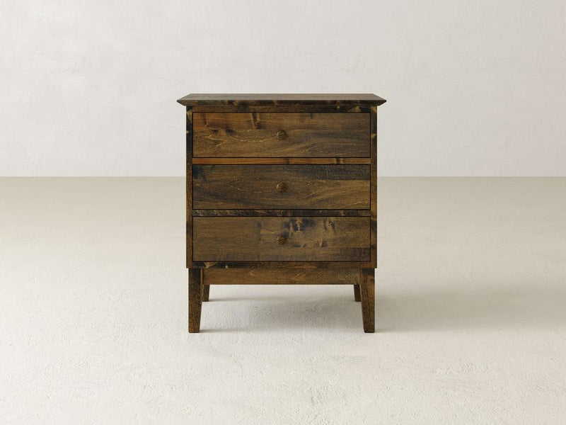 The Bennett 3 Drawer Nightstand by James & James, crafted from solid maple, stands on a light floor against a white wall. It showcases a rustic finish, highlighting the wood grain with simple, clean lines that enhance its traditional design charm.