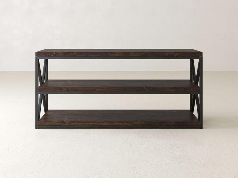 A minimalist dark wood console table from James+James, named the Boyd Shelf, offers three spacious shelves and an industrial steel frame with open sides featuring an X-pattern for added support. The stylish piece is set against a plain, neutral wall and floor.
