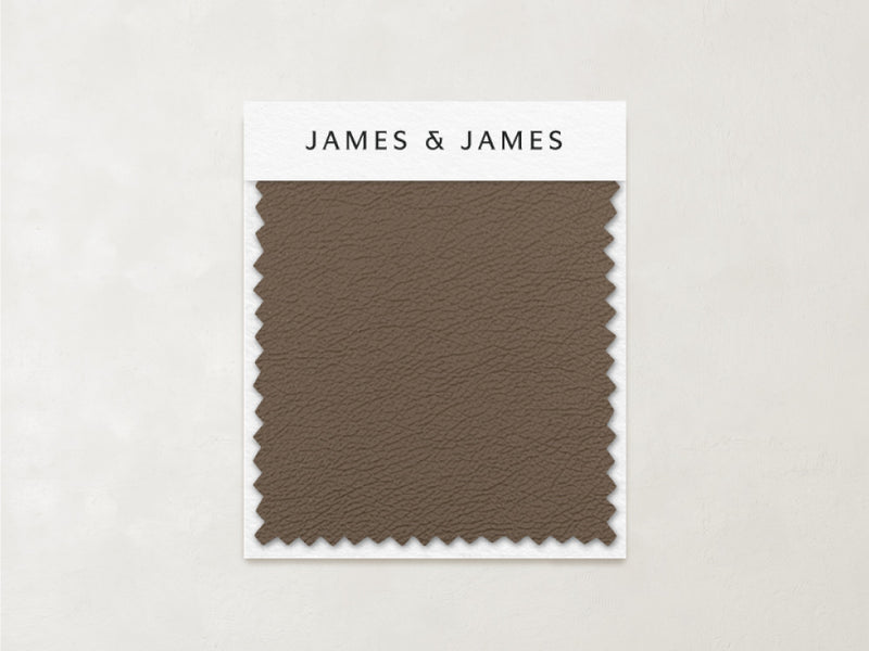 A rectangular sample of cool-toned brown leather, the Toffee - Leather Swatch by James+James, with zigzag-cut edges is attached to a card on a light beige background.