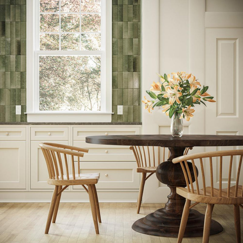 A dining area with a dark wooden round table and three wooden chairs. A vase with orange flowers is on the table. The background features cabinets and a window with a view of trees. The walls have green tile accents.