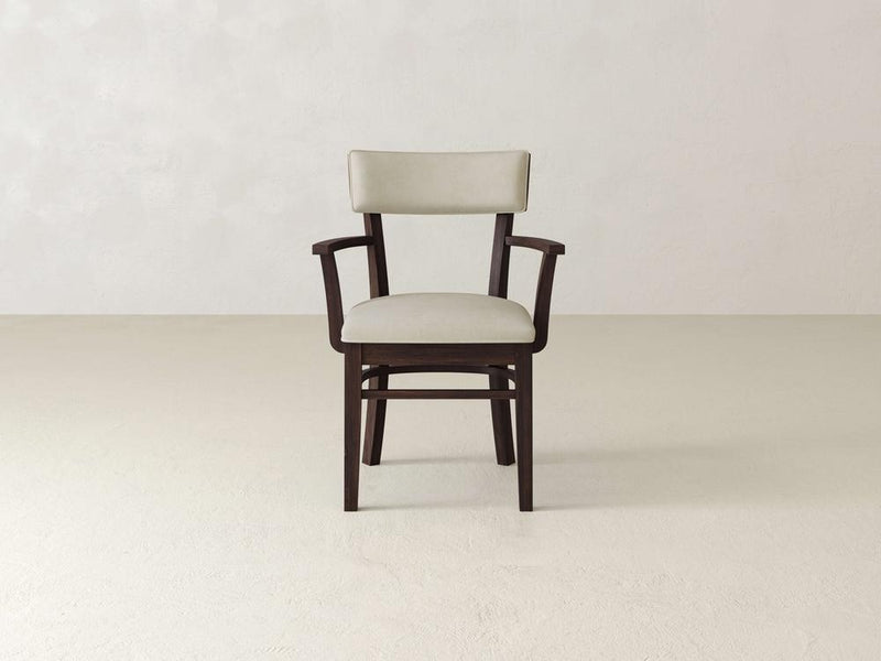 A wooden chair with armrests and a light beige cushioned seat and backrest sits centered in a minimalistic room with off-white walls and floor.