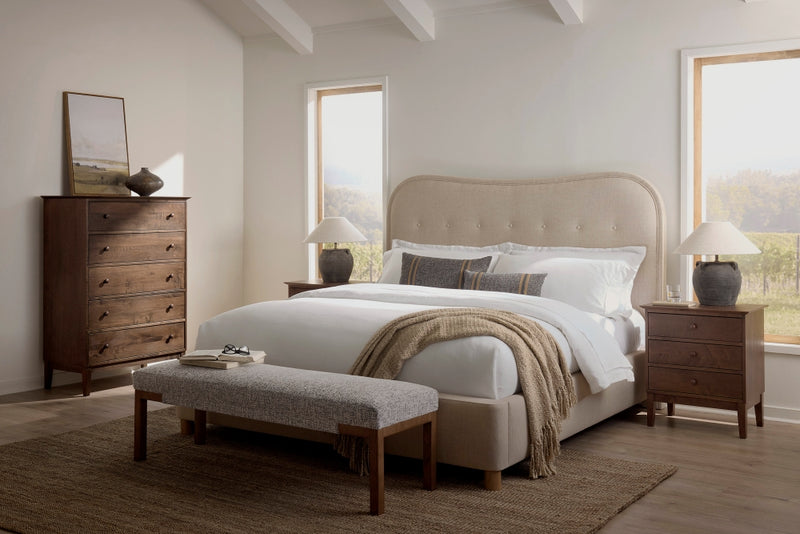 The Florence Bed – Velvet by James & James enhances a cozy bedroom with timeless appeal. Adorned in white bedding and decorative pillows, it’s flanked by wooden nightstands with lamps and complemented by a wood bench at the foot, a tall dresser, and two large windows completing the modern design.