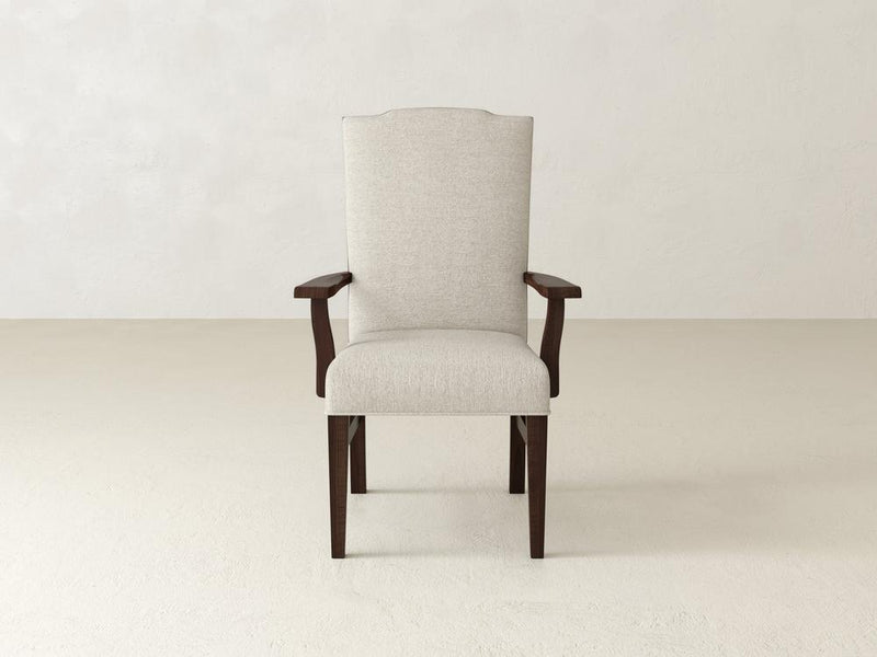 The Hadley Arm Dining Chair - Tobacco by James+James, featuring beige upholstery with wooden armrests and legs, sits on a light-colored floor against a matching plain wall.