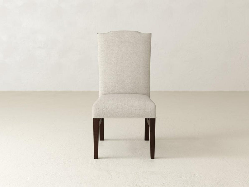 A tall, upholstered dining chair with a cream-colored fabric and dark wooden legs is centered against a light beige, textured wall and floor. The chair has a slightly curved backrest.