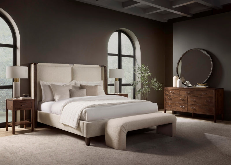 A cozy bedroom showcases the Holden Leather Bed by James & James with cream bedding and an upholstered bench at the foot. Two side tables with lamps flank this solid maple wood bed, while a dresser with a round mirror and a potted plant add decor. Arched windows provide natural light.