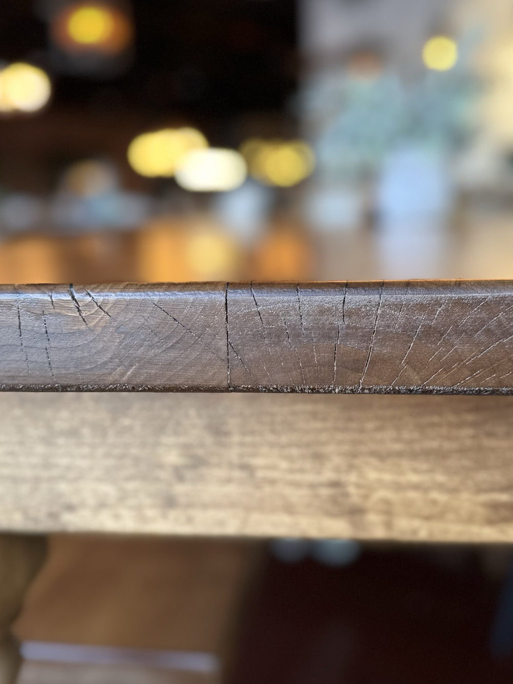 Close-up of a wooden table edge, showing detailed grain and cracks. The background is softly blurred with warm lighting, suggesting an indoor setting, possibly in a cozy room or restaurant.