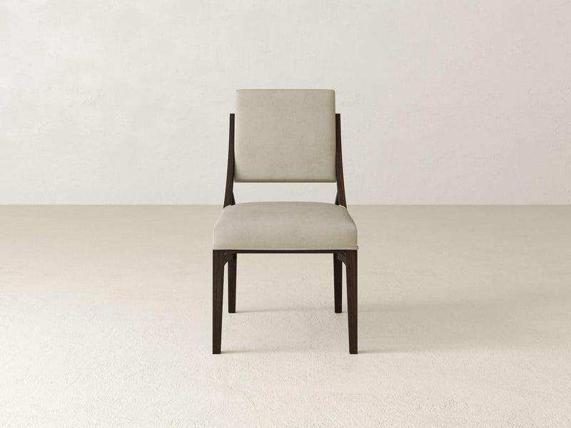 A modern, minimalist chair with a light beige cushioned seat and backrest. The chair features a dark wooden frame and is placed on a light-colored floor with a plain, neutral wall in the background.