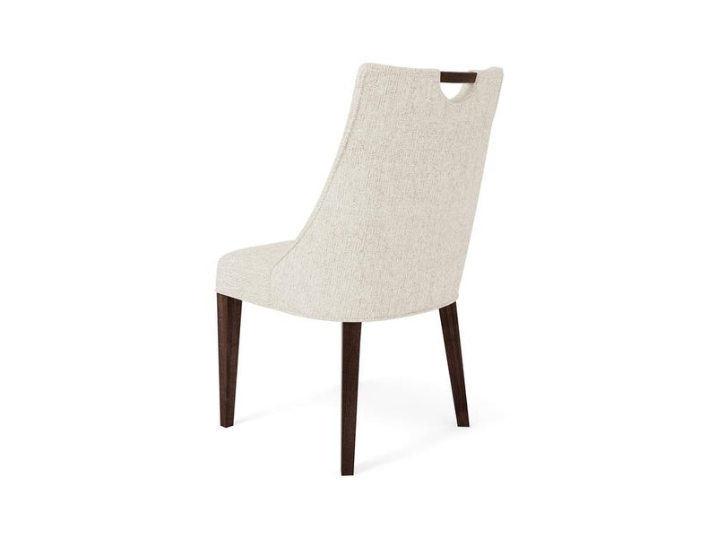 A beige upholstered chair with a tall, slightly curved backrest featuring a small handle cutout at the top. The chair has dark wooden legs and a minimalist design, viewed from a rear-side angle.