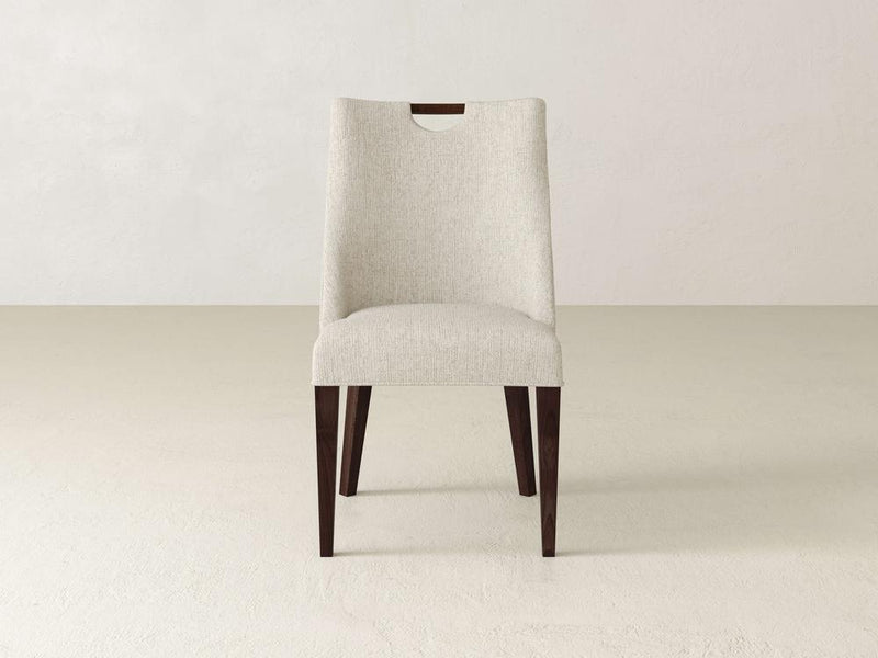 Introducing the Rosalie Dining Chair by James & James, a modern masterpiece in light beige upholstery with a minimalist design, dark wooden legs, and a slightly curved backrest featuring a sleek wooden handle cutout at the top.