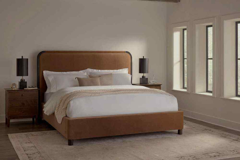 A bedroom exudes modern elegance with the Sloane Bed – Linen by James & James, featuring a brown upholstered headboard and white bedding with a light throw. Flanked by wooden nightstands with black lamps, it sits on a light rug covering the dark floor, while three large windows line the right wall.