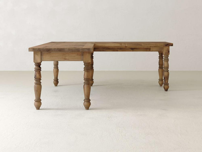 The Turned Leg Corner Desk by James+James, crafted from solid hardwood, features a distinctive L-shaped design and finely carved legs. It stands elegantly on a light-colored floor against a neutral wall, showcasing a natural wood grain finish that enhances its charm as an elegant addition to any workspace.