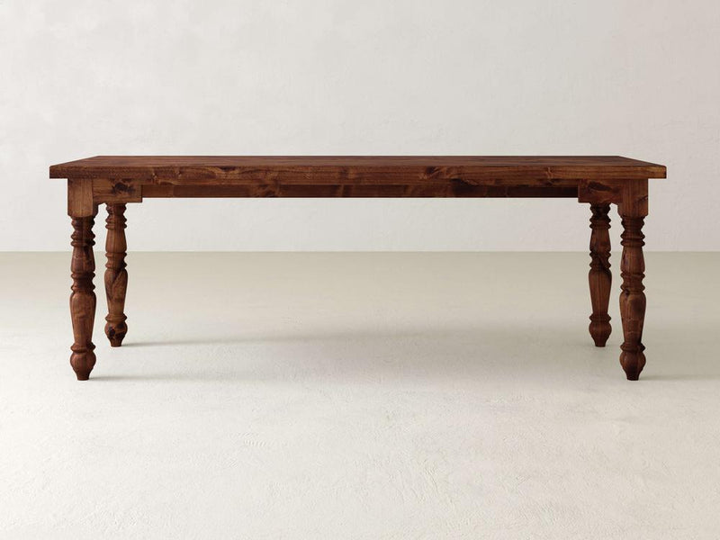 A hardwood dining table with intricately carved legs resembles a Turned Leg Desk by James+James, set elegantly on a light-colored floor against a plain white wall.