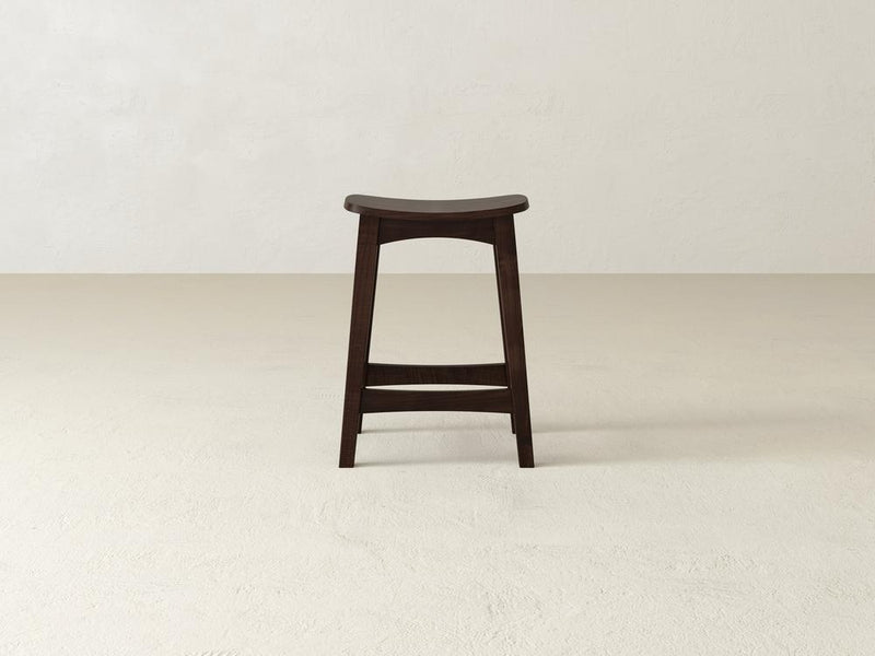 A dark wooden Weston Stool by James & James stands against a plain, light background. Made from durable maple hardwood, it showcases a contoured seat and a minimalist design with four legs and a footrest.
