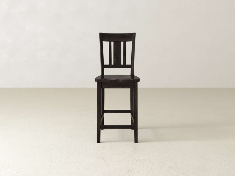 The William Stool by James & James, a handcrafted dark wooden seat, stands alone on a light floor against an off-white wall—ideal for minimalist home decor.