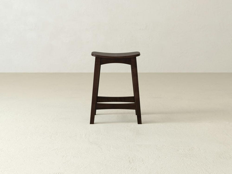 A James & James Weston Stool, made of durable maple hardwood, features a contoured seat. It stands elegantly on a light, textured floor against an off-white wall, highlighting its minimalist design with four sturdy legs.