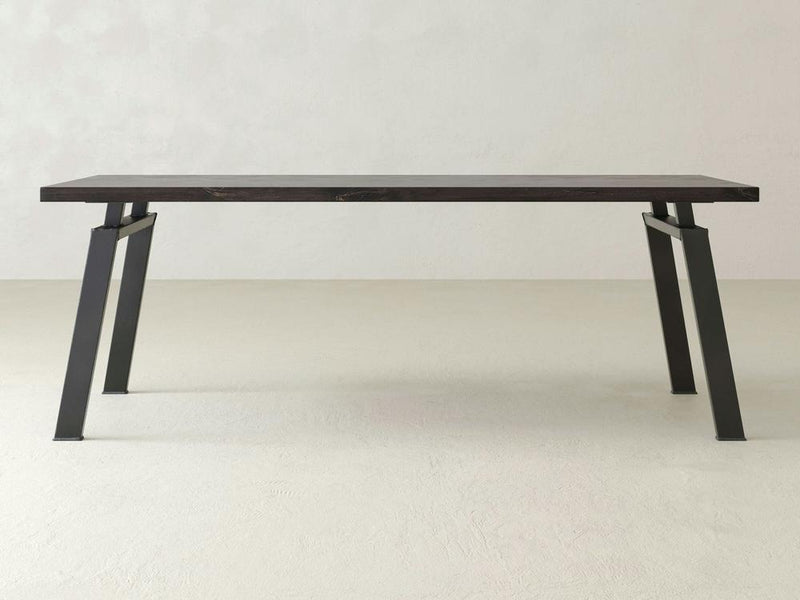 The James+James Fulton Desk, a minimalist wooden table featuring a dark top and angled black metal legs, is showcased in a plain, light-colored room with a concrete floor and textured walls. The design exudes industrial elegance with its sleek lines and sturdy appearance. Customization options are available.