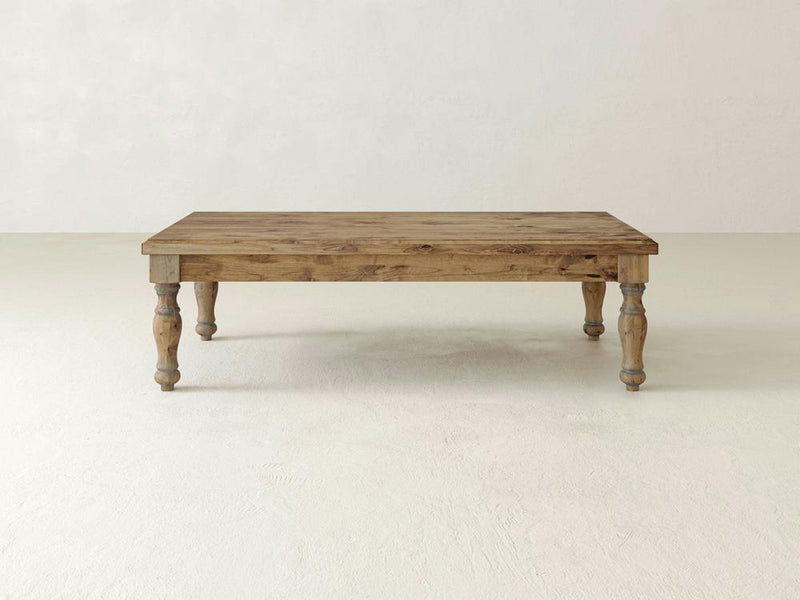 The **Abigail Coffee Table - Harvest Wheat** by **James+James** features a rectangular wooden design with a rustic finish. It stands on four intricately carved legs and boasts a smooth surface that showcases the visible wood grain. The table is placed on an off-white floor against a plain, light-colored background.
