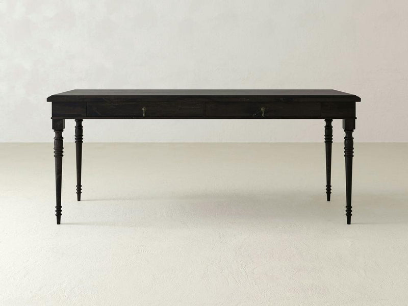 A striking Audrey Desk - Charred Ember from James+James, showcasing a dark wooden finish and turned legs, with two small drawers on either side. Set against a plain light-colored background, this desk embodies a simplistic and classic design while standing confidently on a flat surface.