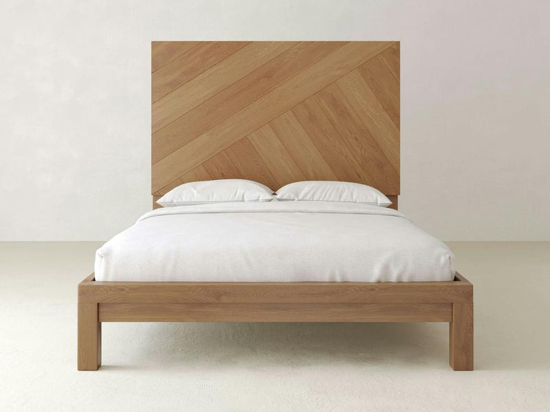 Introducing the Luna Bed by James+James: a minimalist masterpiece made from natural white oak, showcasing a tall headboard adorned with an elegant chevron pattern. The bed is perfectly complemented by crisp white bedding and pillows, set against light-colored walls and flooring that beautifully accentuate the natural wood finish of this refined piece.