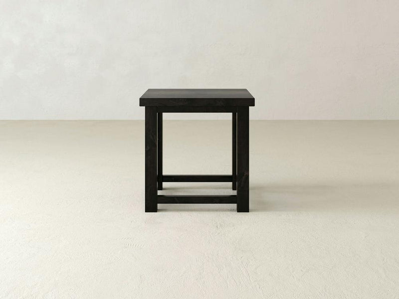The Carter X Side Table - Charred Ember from James+James stands against a plain off-white backdrop. This minimalist black wooden piece features a rectangular top with simple, straight legs and a basic support structure, embodying a clean and modern design aesthetic.