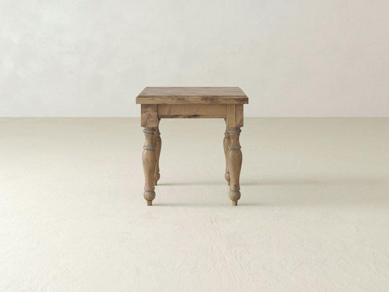 An Abigail End Table - Harvest Wheat by James+James with a natural finish and a rustic design is centered in the image. The table features four carved legs and a simple rectangular top. The background showcases a minimalistic, off-white room with smooth walls and floor.