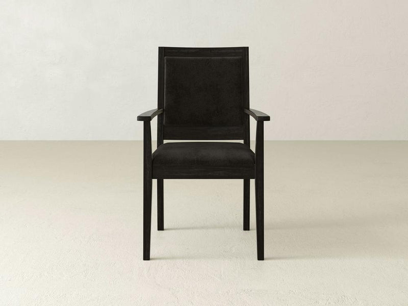 The Gina Arm Dining Chair by James+James boasts a contemporary silhouette, crafted from wood with dark-colored upholstery set against a plain, light-colored backdrop. Featuring armrests and a straight, rectangular backrest, this chair's minimalist design exudes modern elegance.