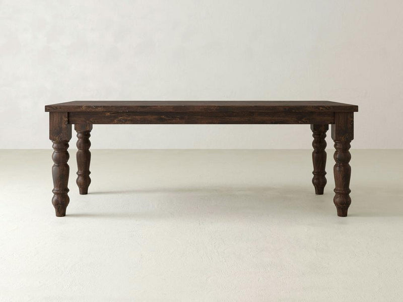 The Baluster Dining Table - Tobacco by James+James is a dark brown wooden dining table with an ornate design, featuring four intricately carved legs and a rectangular top. The background is a minimalist, light-colored wall and floor.