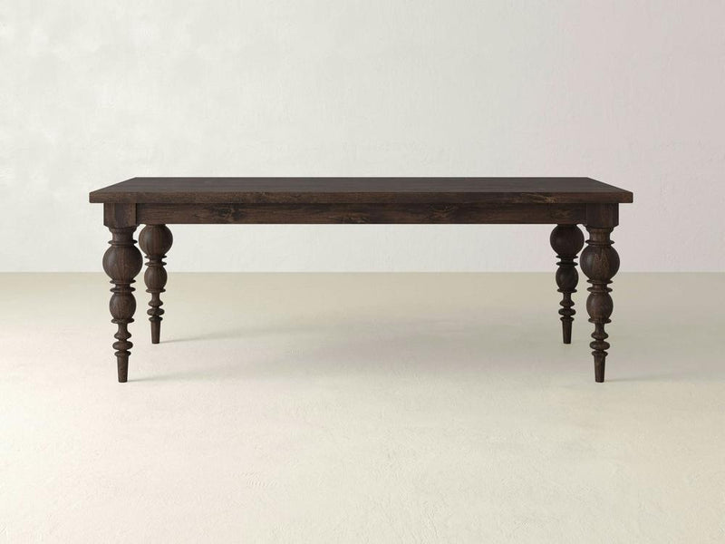 The Bailey Dining Table - Tobacco by James+James, with intricately carved, turned legs and a dark wooden finish, stands on a light-colored floor against a plain, light gray wall. The rectangular tabletop features a polished surface that beautifully showcases the natural wood grain.