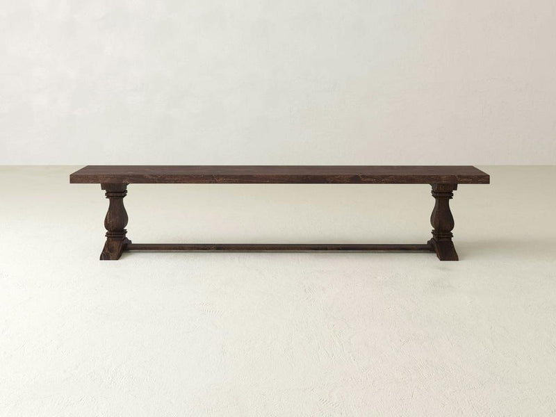 Introducing the Heirloom Bench by James+James: a long, rectangular wooden masterpiece with a dark brown finish. This elegant bench features two meticulously carved legs on each end, connected by a horizontal support bar, exuding timeless sophistication against a plain, light-colored background.