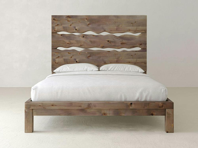 A James+James Live Edge Bed with a simple, rustic design sits in a minimalistic room. The handcrafted headboard features two wavy cut-out patterns, enhancing its unique artistic appeal. The bed is adorned with white bedding, including pillows and a neatly tucked duvet. The background is plain and light-colored.