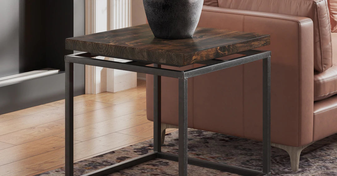 A stylish wooden end table with black metal legs sits next to a beige leather sofa. The table is adorned with a large, dark ceramic vase. The room has hardwood floors and a patterned rug.