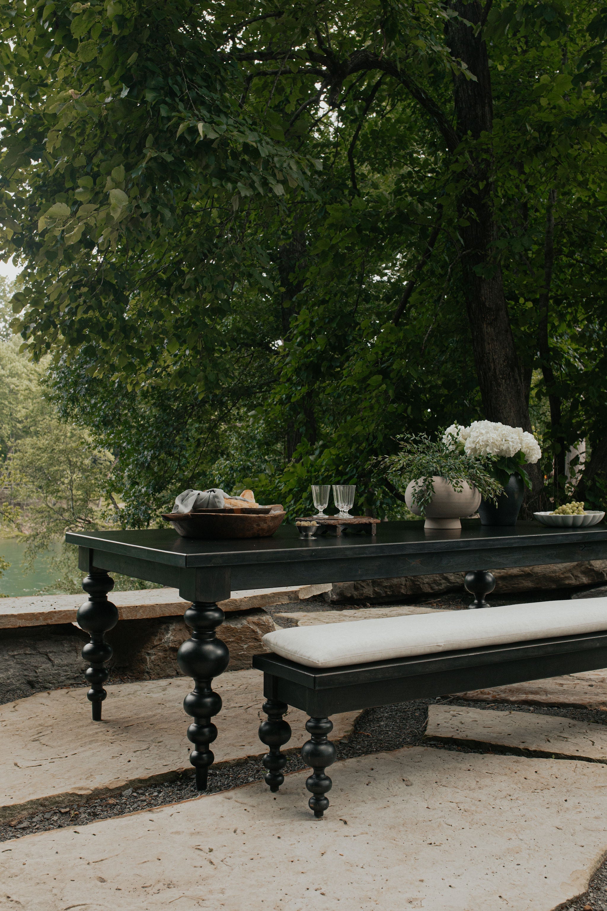 A dark wooden outdoor table with a cushioned bench is set with flowers, glasses, and bread, surrounded by lush green trees and a stone patio near a peaceful body of water.