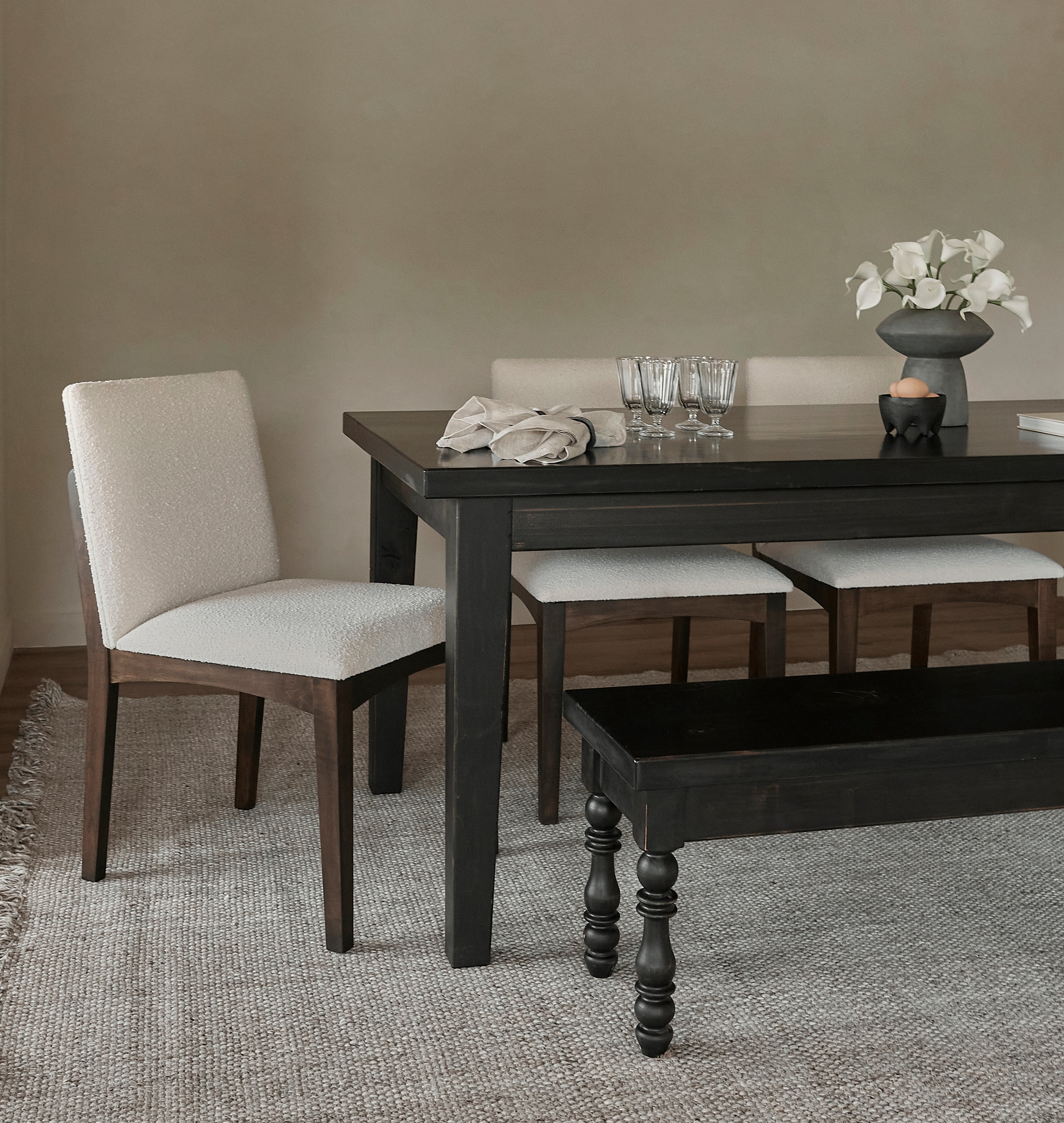 A cream-colored, upholstered dining chair with dark wooden legs sits next to a dark wooden table on a textured beige rug in a minimalist room.