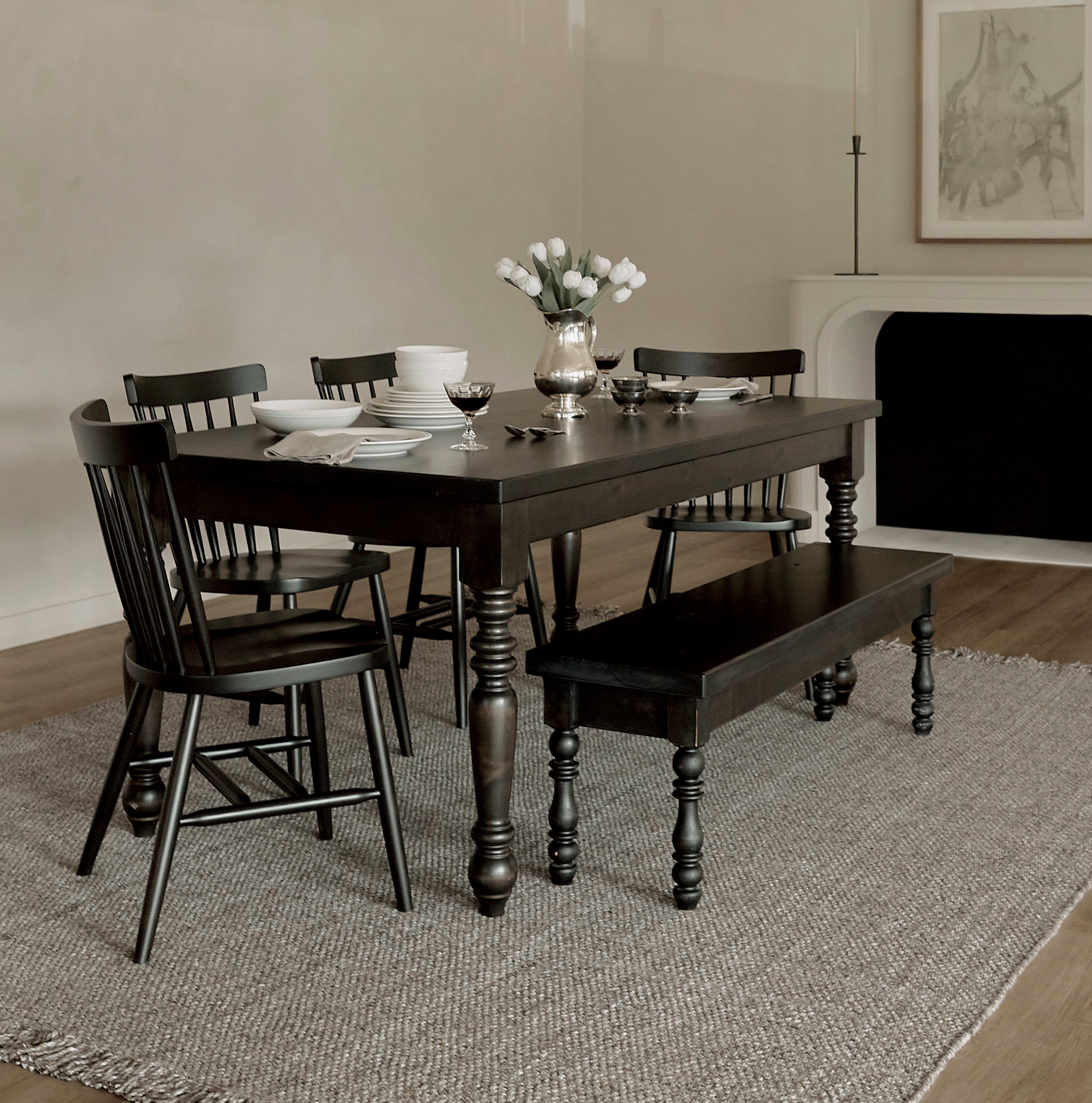 A dining room with a dark wooden table, four chairs, and a bench on a textured rug. A silver vase with white tulips is on the table, and a modern chandelier hangs above. Art and a candlestick are in the background.