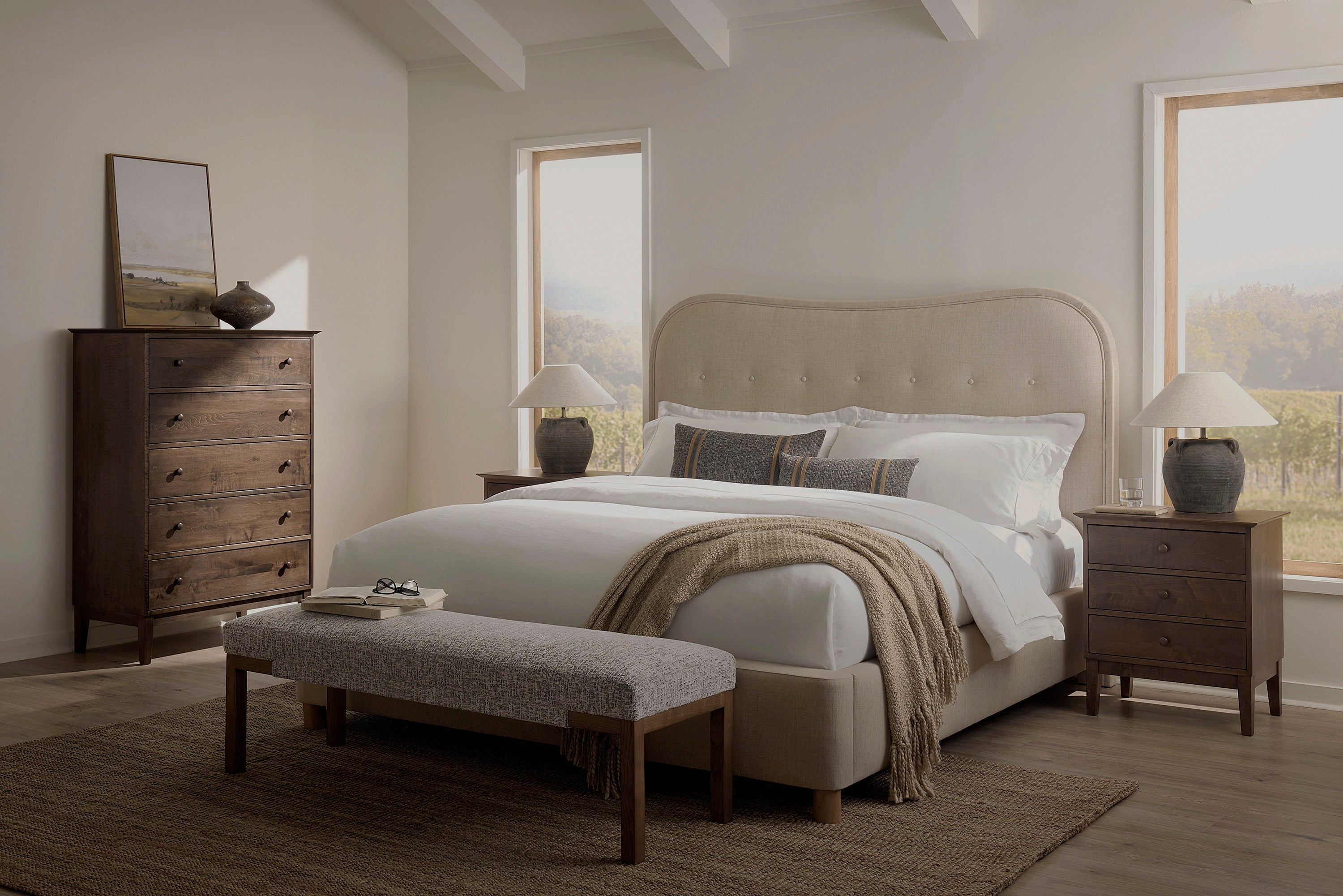 A modern bedroom with a beige upholstered bed, white bedding, two wooden nightstands with lamps, a chest of drawers, a landscape painting, a gray bench, and large windows with scenic views.